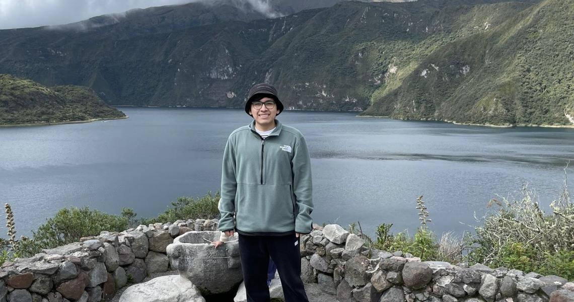A young Ecuadorian man in a periwinkle blue fleece Northface jacket, black slacks and grey bucket hat smiles at the camera, standing infront of a knee high cobblestone wall and primitive looking rock vase that seperate him from a large rippling lake and steep brush-covered mountains in the background.