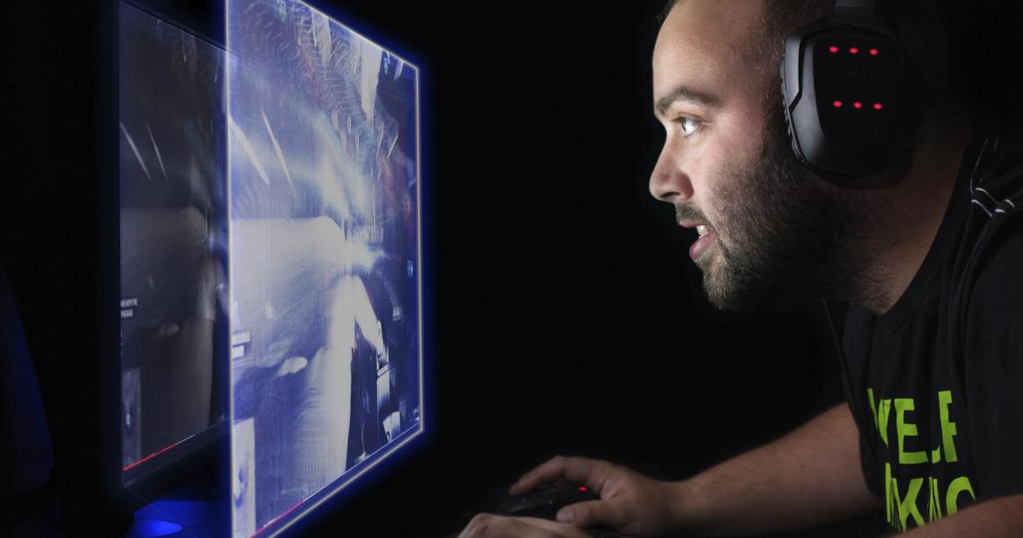 The profile of a late 20s bearded white mans face is illuminated by a computer screen which hes leaning toward. He wears a gaming headset with red LEDs and is using a mouse to play a shooter video game. Projected from the computer screen is a second image of the display, like a larger hologram of the screen he is playing on.