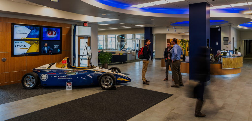 Three men meet in the lobby of Notre Dame's Idea Center while a woman passes through; a promotional F1 car sits to the side, a symbol of the Center’s race car-related branding