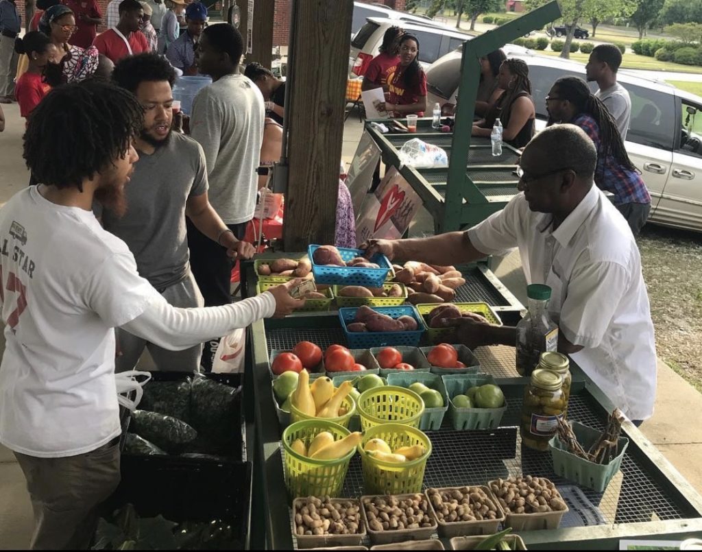 Two Tuskegee students purchase a small basket of sweet potatoes from a local vendor as the bustle of a monthly farmer's market carries on in the background. An assortment of produce is displayed on the table between them, vibrant colors gleaming through the shade of the market's awning.