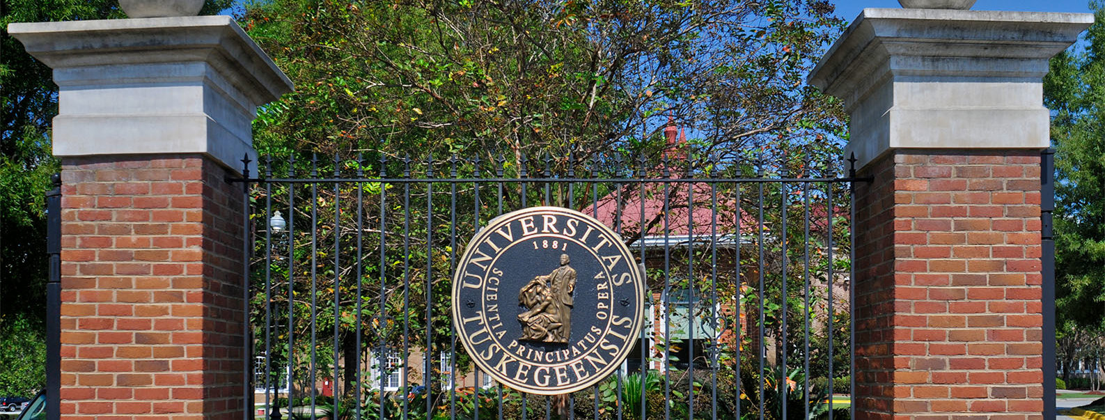 Two free standing square brick pillars rise a dozen feet from a base of shrubbery. A wrought iron fence supporting the circular seal of Tuskegee University emblazoned with the latin text for "Knowledge, Leadership, Service" is in between along with a depiction of the campus statue dedicated to Booker T. Washington, then Tuskegee Institutes's founding president. Foliage, roads and campus buildings occupy the background.