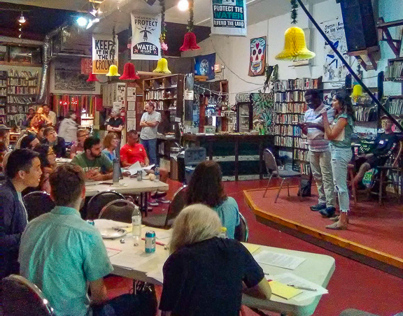 Two students present questions of sustainability to a small crowd in an eclectically decorated community center. They stand atop a step-high elevated stage surrounded by stocked wooden bookcases, environmental posters and strings of fairy lights all towering over a dark crimson floor.