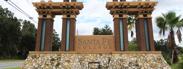 Atop a raised cobblestone base sits a two-tier pyramid of brick arches, each topped with a lattice of wooden cross beams. Between the lower two arches is a clay colored slab reading Santa Fe College, while the top arch supports the S.F. initials in gold and black.