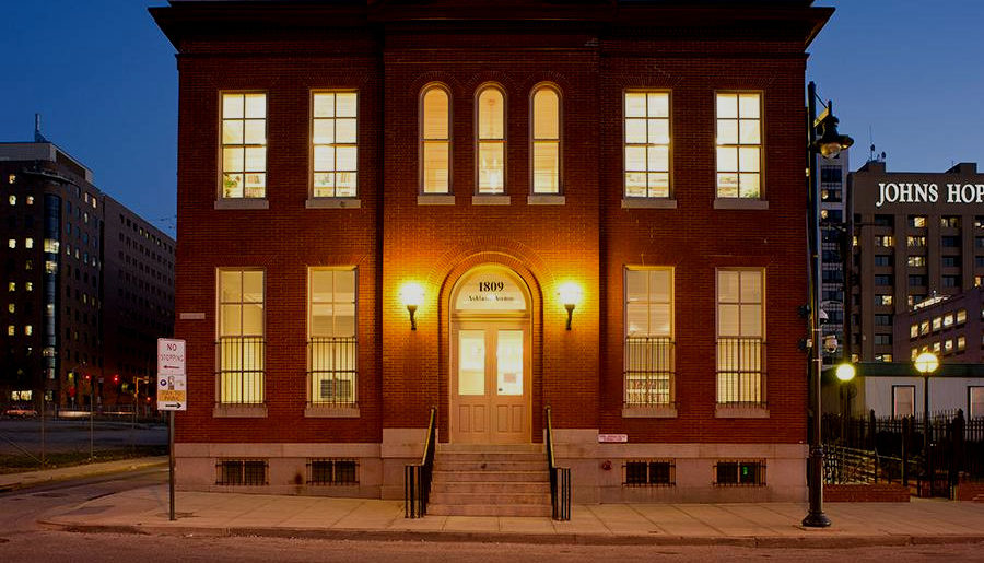 The darkness of dusk falls over a New England colonial style, two level red brick building which occupies the center of the image; Deering Hall. The building's face is lit by two incandescent sconces bordering a white wood and glass door atop a small porch staircase. A symmetrical collection of windows adorns the building's face, contrasting the shadowed city scape in the background.