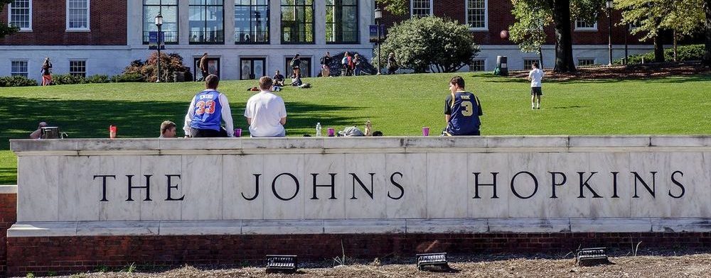 A stout, wide marble sign stretches across the lower half of the image, adorned in thin, black serif font with "THE JOHNS HOPKINS." Atop the sign sit three young men next to their water bottles and backpacks, looking on a neatly mowed green quad speckled with students and shrubbery. A brick and marble building fills the backdrop.