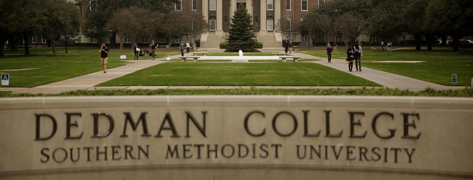 The name of Southern Methodist University's Dedman College juts out in bold, black serif letters from a beige stone sign spanning the lower third of the shot. Above it, in the near distance, stands the marble columns, red brick walls and copper-green dome of Dallas Hall, distanced by concrete walkways cutting across a neatly trimmed campus quad
