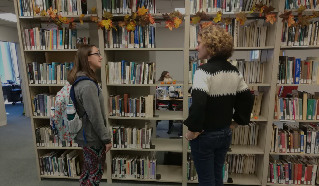 Two women stand in front of an off white metal bookshelp filled with colorful covers. A strand of autumnal leaves hangs along the top of the bookshelf, and a small gap on one of the two dozen shelf hosts a small collection of pumpkins, indicating the large modern library space is celebrating the Fall season.