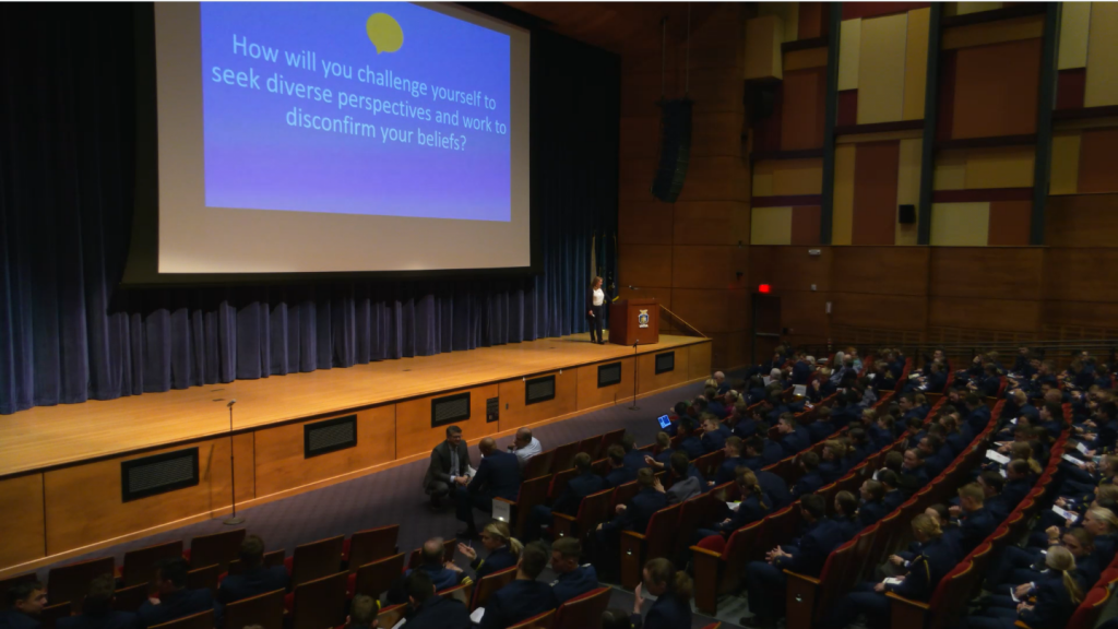 A large wood panelled auditorium is filled with Coast Guard cadets, sat in the curved rows of auditorium folding seats to watch a presentation on a massive pull down presentation screen. A women stands beside a decorated wooden lectern, delivering the presentation.